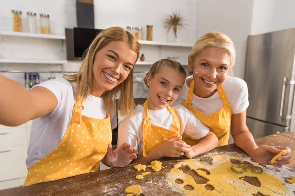 Щаслива дитина з матір'ю і бабусею посміхається на камеру, готуючи печиво разом на кухні — стокове фото