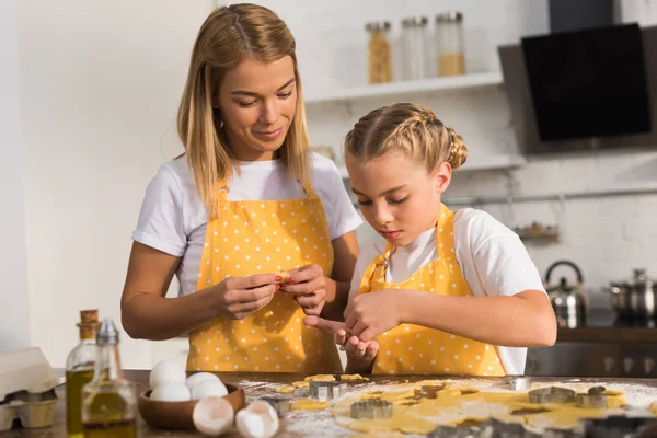 Beautiful mother and daughter in aprons preparing cookies together in kitchen — Stock Photo