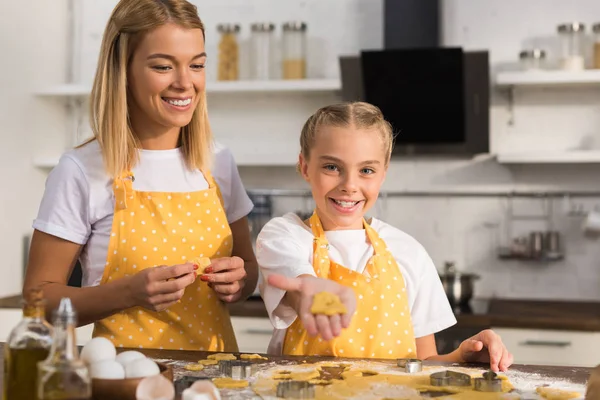 Happy mother and daughter in aprons holding unbaked cookies in kitchen — Stock Photo