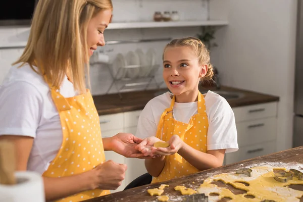 Счастливые мать и дочь в фартуках улыбаются друг другу, пока готовят печенье вместе — стоковое фото