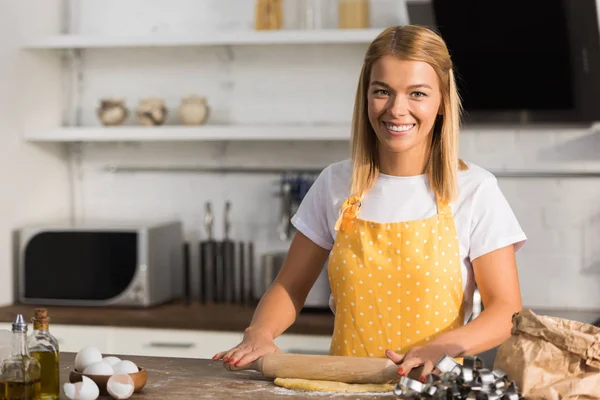 Felice giovane donna con mattarello preparare pasta e sorridere alla macchina fotografica in cucina — Foto stock