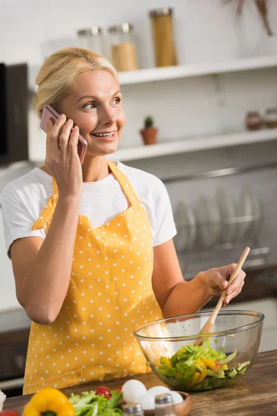 Sonriente mujer madura en delantal hablando por teléfono inteligente y mirando hacia otro lado mientras cocina ensalada de verduras en la cocina - foto de stock