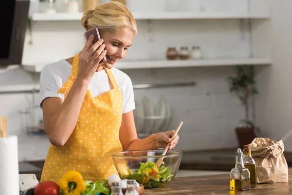 Smiling mature woman in apron talking by smartphone while cooking in kitchen — Stock Photo