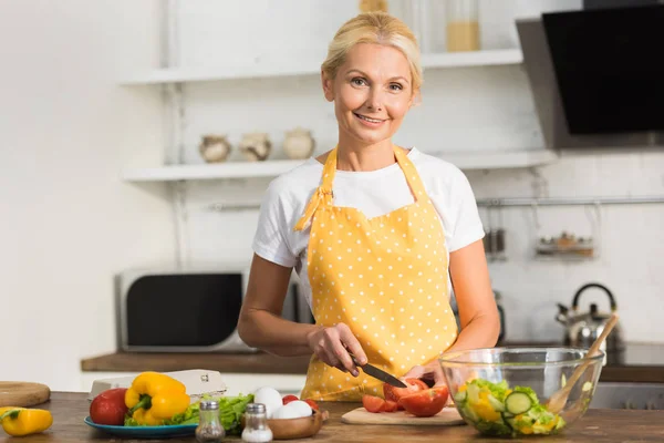 Heureuse femme mûre dans tablier souriant à la caméra tout en cuisinant dans la cuisine — Photo de stock