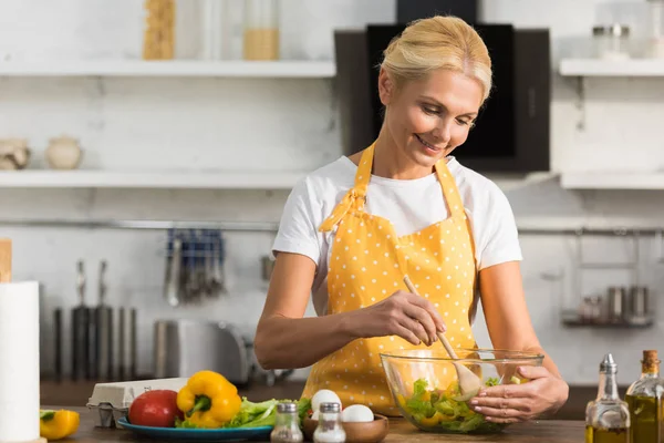 Happy mature woman in apron cooking vegetable salad in kitchen — Stock Photo