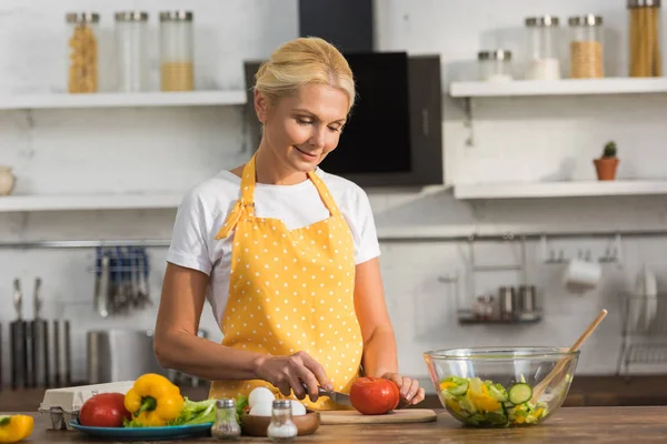 Sonriente mujer madura en delantal cocina ensalada de verduras en la cocina - foto de stock