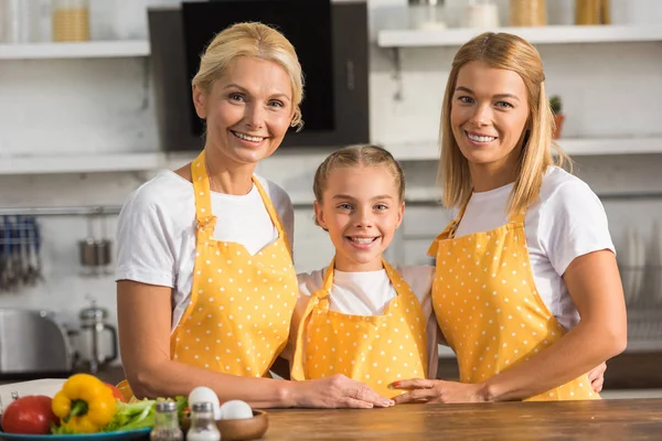 Feliz familia de tres generaciones en delantales sonriendo a la cámara en la cocina - foto de stock