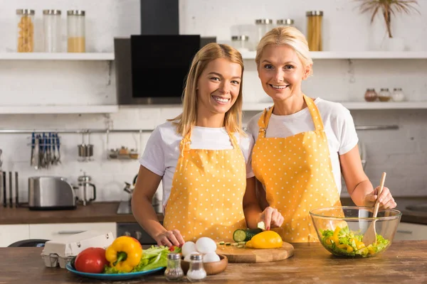 Feliz madre adulta e hija en delantales sonriendo a la cámara mientras cocinan juntos - foto de stock