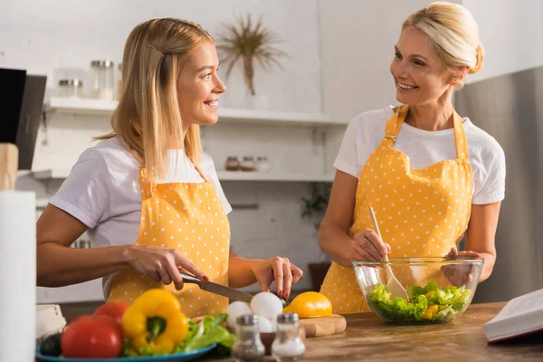 Happy mature woman and young daughter smiling each other while cooking vegetable salad together — Stock Photo