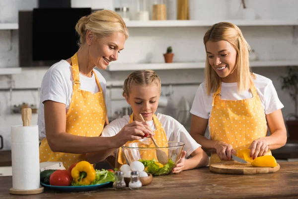Happy child with mother and grandmother cooking vegetable salad together in kitchen — Stock Photo
