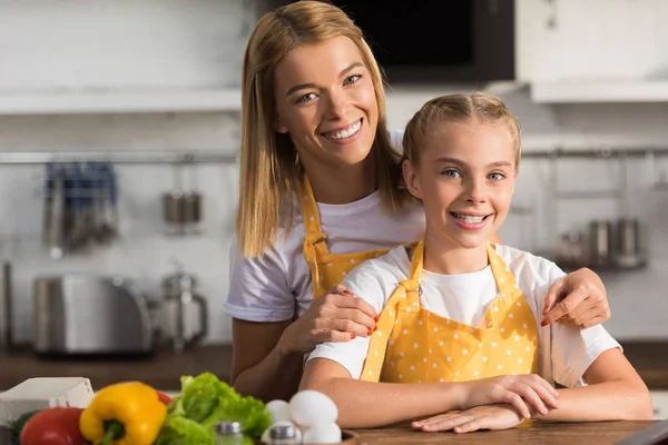 Hermosa feliz madre e hija en delantales sonriendo a la cámara en la cocina - foto de stock