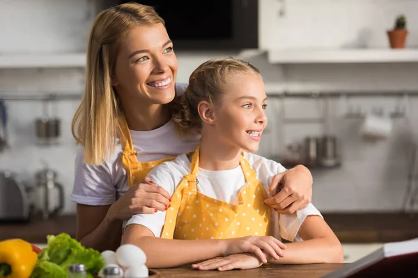 Glückliche Mutter und Tochter in Schürzen lächelnd und wegschauend in der Küche — Stockfoto