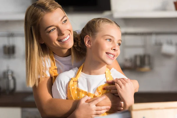 Feliz madre e hija en delantales abrazándose y mirando hacia otro lado en la cocina - foto de stock