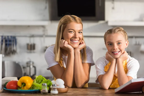 Feliz madre e hija en delantales apoyados en la mesa de la cocina y sonriendo a la cámara - foto de stock