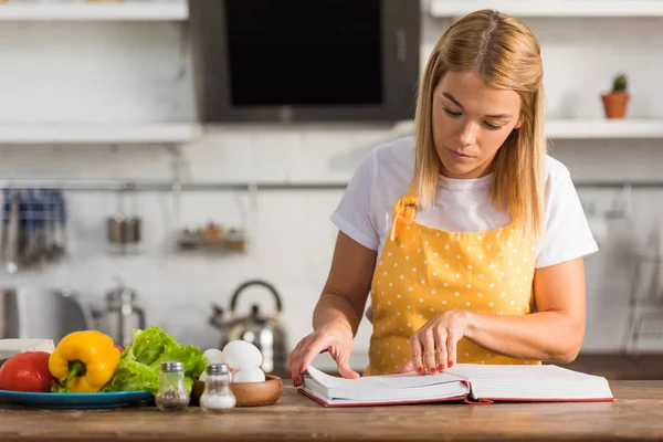 Mujer joven en delantal leyendo libro de cocina mientras cocina en la cocina - foto de stock