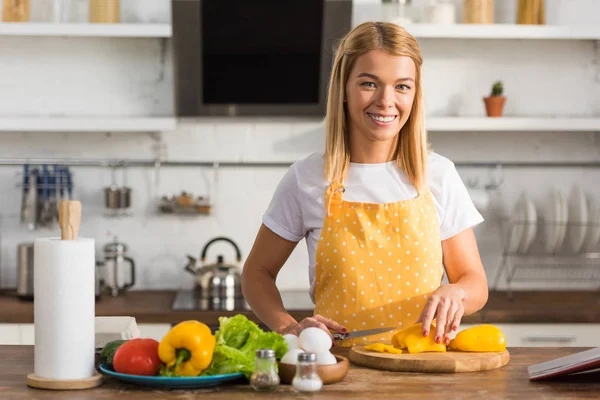 Felice giovane donna che taglia verdure e sorride alla macchina fotografica in cucina — Foto stock