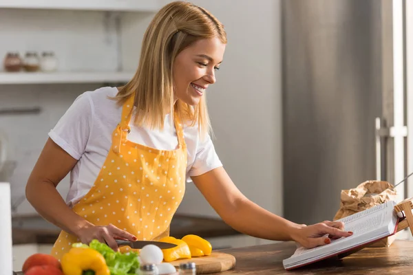 Sorridente giovane donna in grembiule leggere libro di cucina mentre cucina in cucina — Foto stock