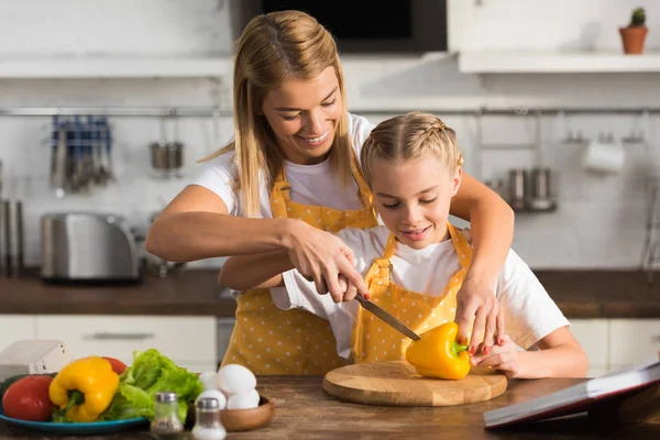 Sorrindo jovem mulher ajudando pequena filha a cortar pimenta na cozinha — Fotografia de Stock