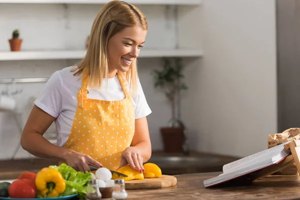 Smiling young woman in apron cutting vegetables and reading cookbook in kitchen — Stock Photo