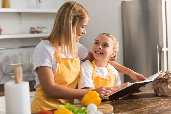 Красивая счастливая мать и дочь улыбаются друг другу и читают кулинарную книгу на кухне — стоковое фото