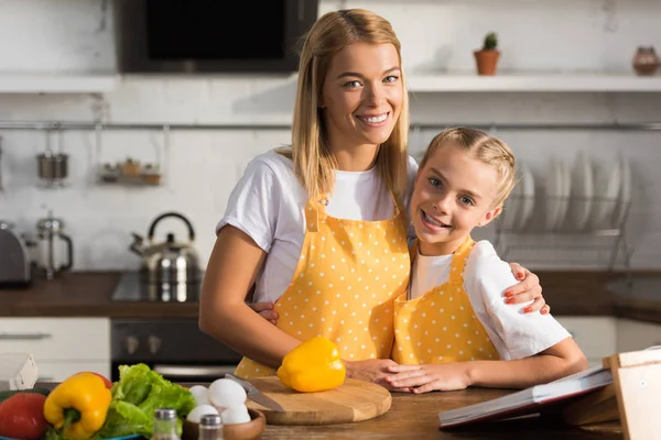 Hermosa feliz madre e hija en delantales sonriendo a la cámara mientras cocinan juntos - foto de stock