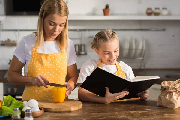 Giovane madre taglio pepe e sorridente figlia lettura ricettario — Foto stock