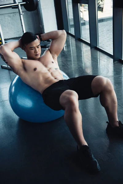Enfocado joven asiático deportista con desnudo pecho haciendo abdominales ejercicio en fitness pelota en gimnasio - foto de stock