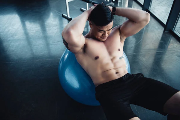 Focado jovem asiático desportista com peito nu fazendo abs exercício no fitness bola no ginásio — Fotografia de Stock