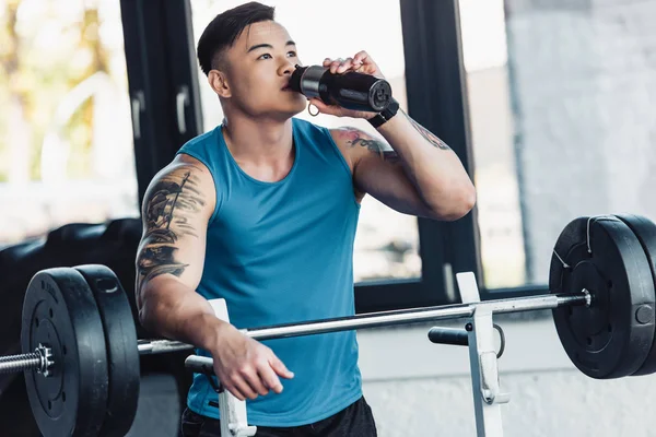 Muscular asian sportsman drinking water and training with barbell at gym — Stock Photo