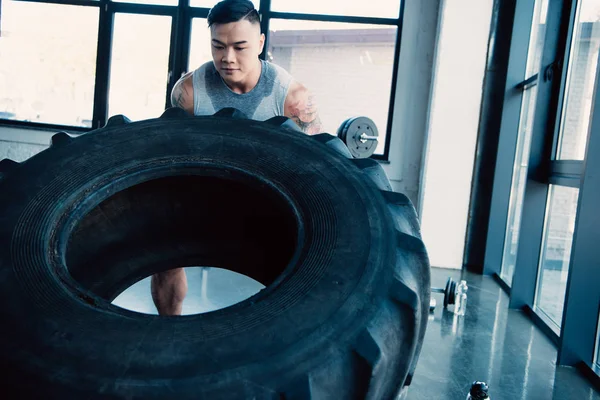 Концентрированный молодой спортсмен прокатывает тяжелую шину в спортзале — стоковое фото