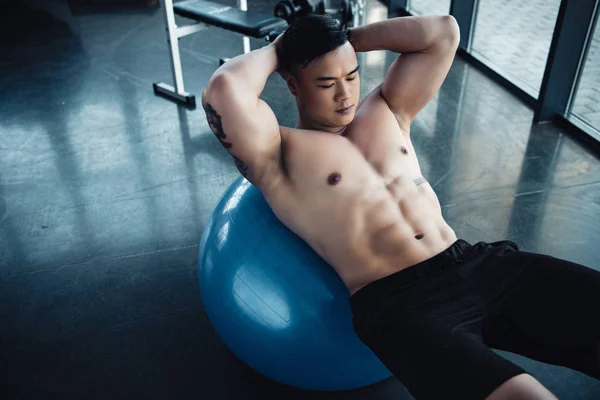 Joven deportista con el pecho desnudo haciendo ejercicios abdominales en la pelota de fitness en el gimnasio - foto de stock