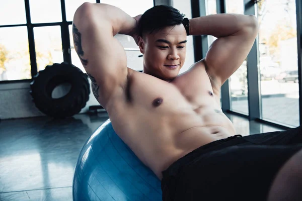 Guapo joven asiático deportista con desnudo pecho haciendo abdominales ejercicio en fitness pelota en gimnasio - foto de stock