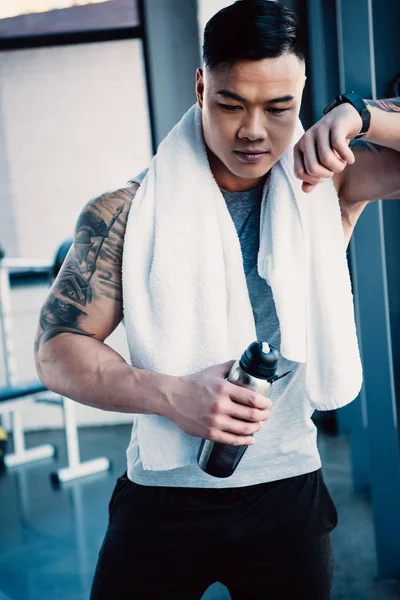 Молодой уставший азиатский спортсмен с полотенцем и бутылкой воды после долгой тренировки — стоковое фото