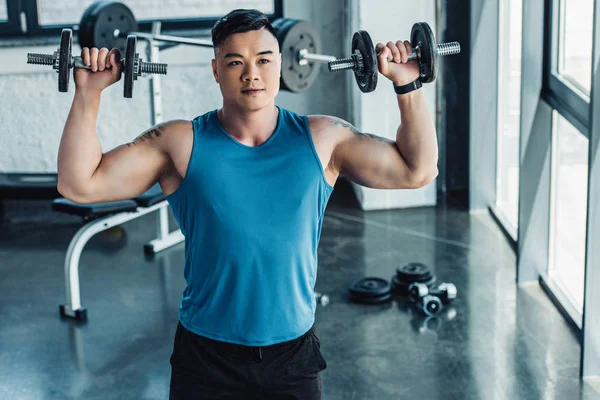 Concentrated young sportsman exercising with dumbbells in gym and looking at camera — Stock Photo