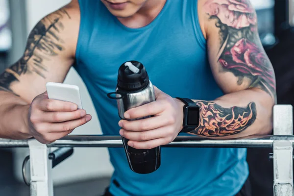 Cropped view of sportsman using smartphone and holding metallic bottle of water — Stock Photo