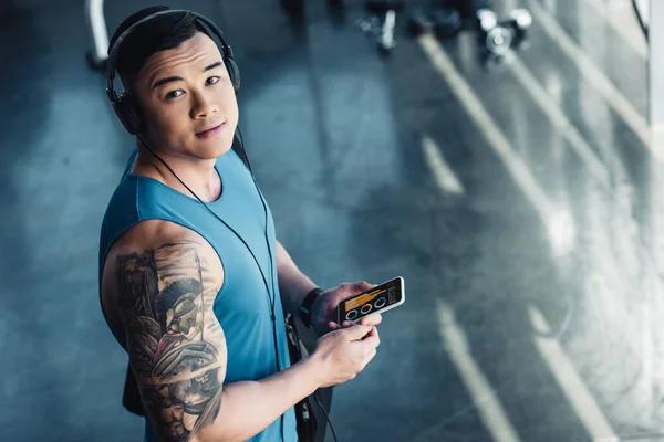 Молодой азиатский спортсмен, использующий смартфон с инфографикой на экране и слушающий музыку — стоковое фото
