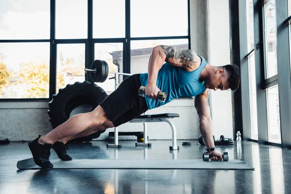Vista lateral del joven deportista haciendo ejercicio con pesas en el gimnasio en la alfombra deportiva - foto de stock