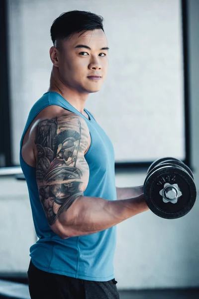 Joven tatuado asiático deportista ejercitando con dumbbell en gimnasio y mirando cámara - foto de stock