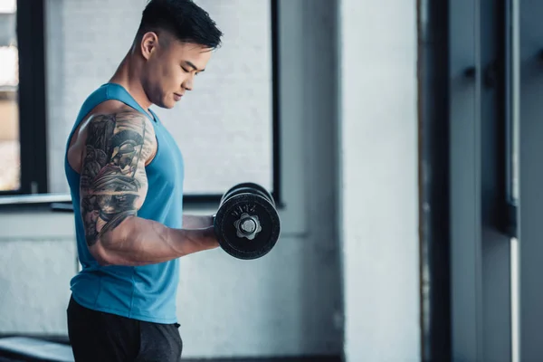 Vista lateral concentrada de un joven deportista haciendo ejercicio con mancuerna en el gimnasio - foto de stock