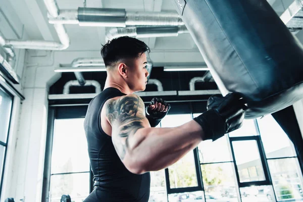 Вид сбоку на молодого азиатского спортсмена в боксёрских перчатках и боксерской сумке в спортзале — стоковое фото