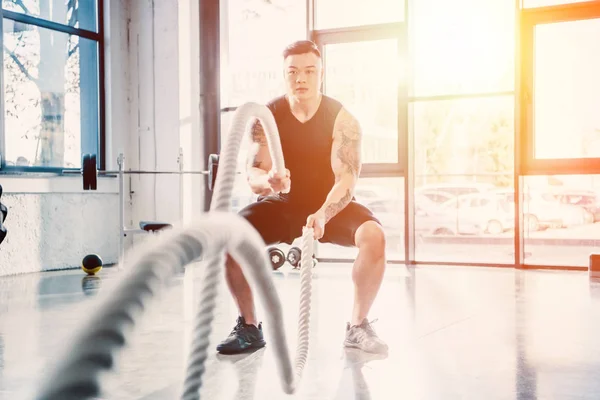 Young sportsman working out with battle ropes at gym in sunlight — Stock Photo