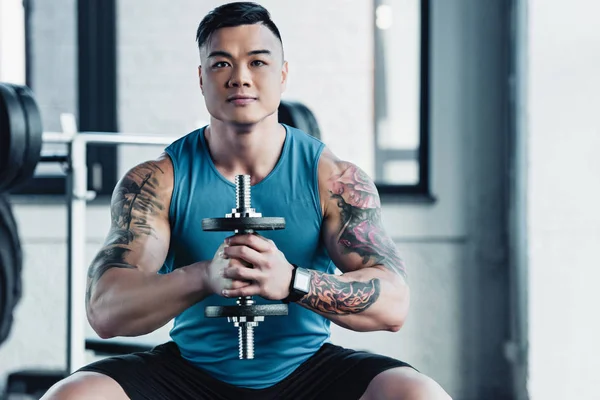 Concentrado joven asiático deportista ejercitando con dumbbell en gimnasio y mirando cámara - foto de stock