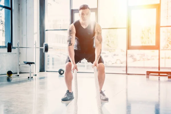 Young sportsman working out with battle ropes at gym in sunlight — Stock Photo