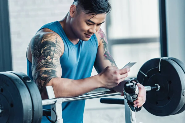 Jeune sportif tatoué souriant utilisant un smartphone dans la salle de gym — Photo de stock