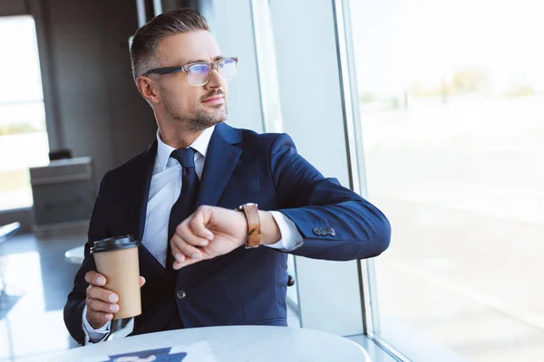 Homem de negócios bonito em óculos com café para ir e relógios olhando para a janela no aeroporto — Fotografia de Stock