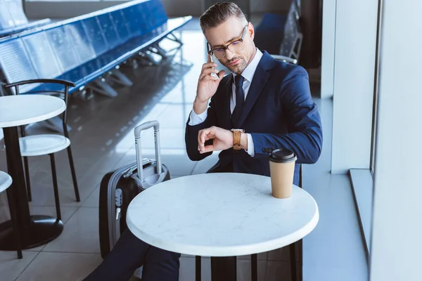 Взрослый бизнесмен смотрит на часы и разговаривает на смартфоне в аэропорту — стоковое фото
