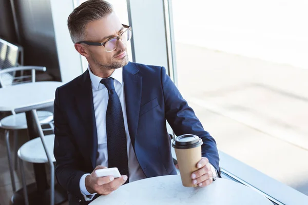 Adult handsome businessman in glasses using smartphone and looking at window at airport — Stock Photo