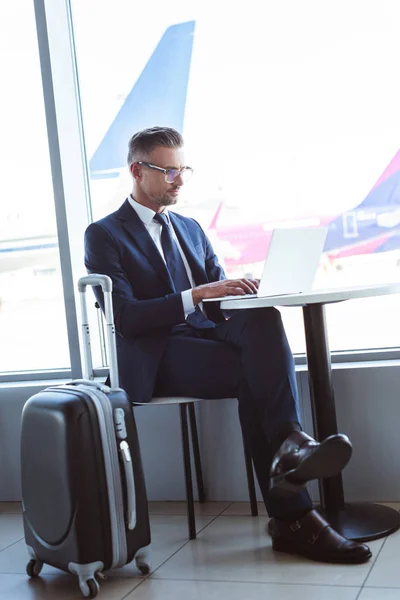 Взрослый бизнесмен в очках, печатающий на ноутбуке за столом в аэропорту — стоковое фото