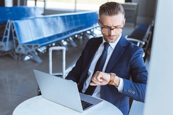 Businessman in glasses using laptop and looking at watch in airport — Stock Photo