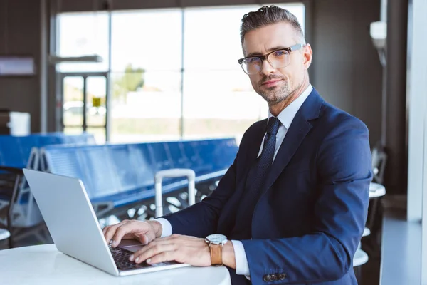 Schöner Geschäftsmann mit Brille blickt in die Kamera und benutzt Laptop am Flughafen — Stockfoto